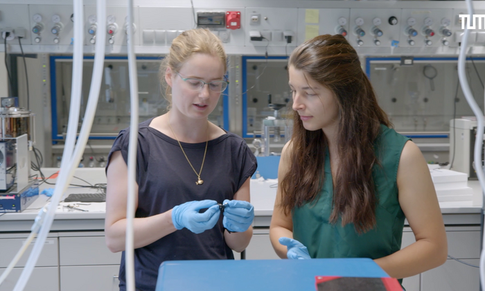 Die Physik-Studentin Nina Miller steht in einem Labor und lässt sich von einer Forschenden einen wissenschaftlichen Sachverhalt erklären.