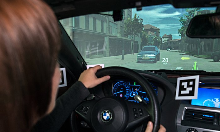 Eine Frau sitzt im Fahrzeugsimulator und blickt auf die Leinwand, auf der das Head-up-Display zu sehen ist.