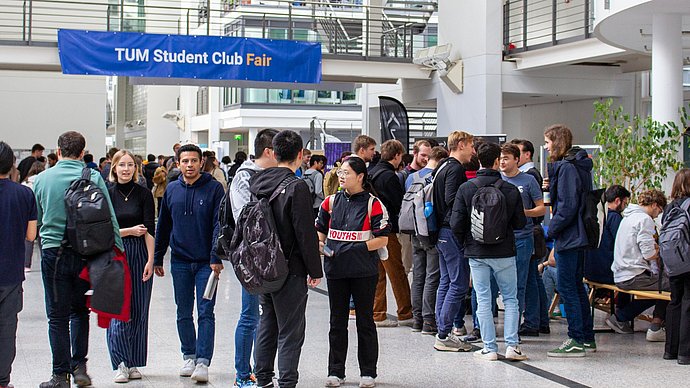 Impression of the TUM Student Club Fair at the Garching campus: Students in the main hall of the Mechanical Engineering building, at a crossing a blue banner with the inscription “TUM Student Club Fair”