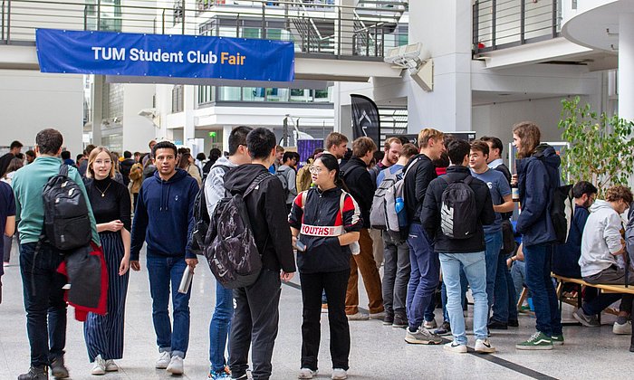 Impression of the TUM Student Club Fair at the Garching campus: Students in the main hall of the Mechanical Engineering building, at a crossing a blue banner with the inscription “TUM Student Club Fair”