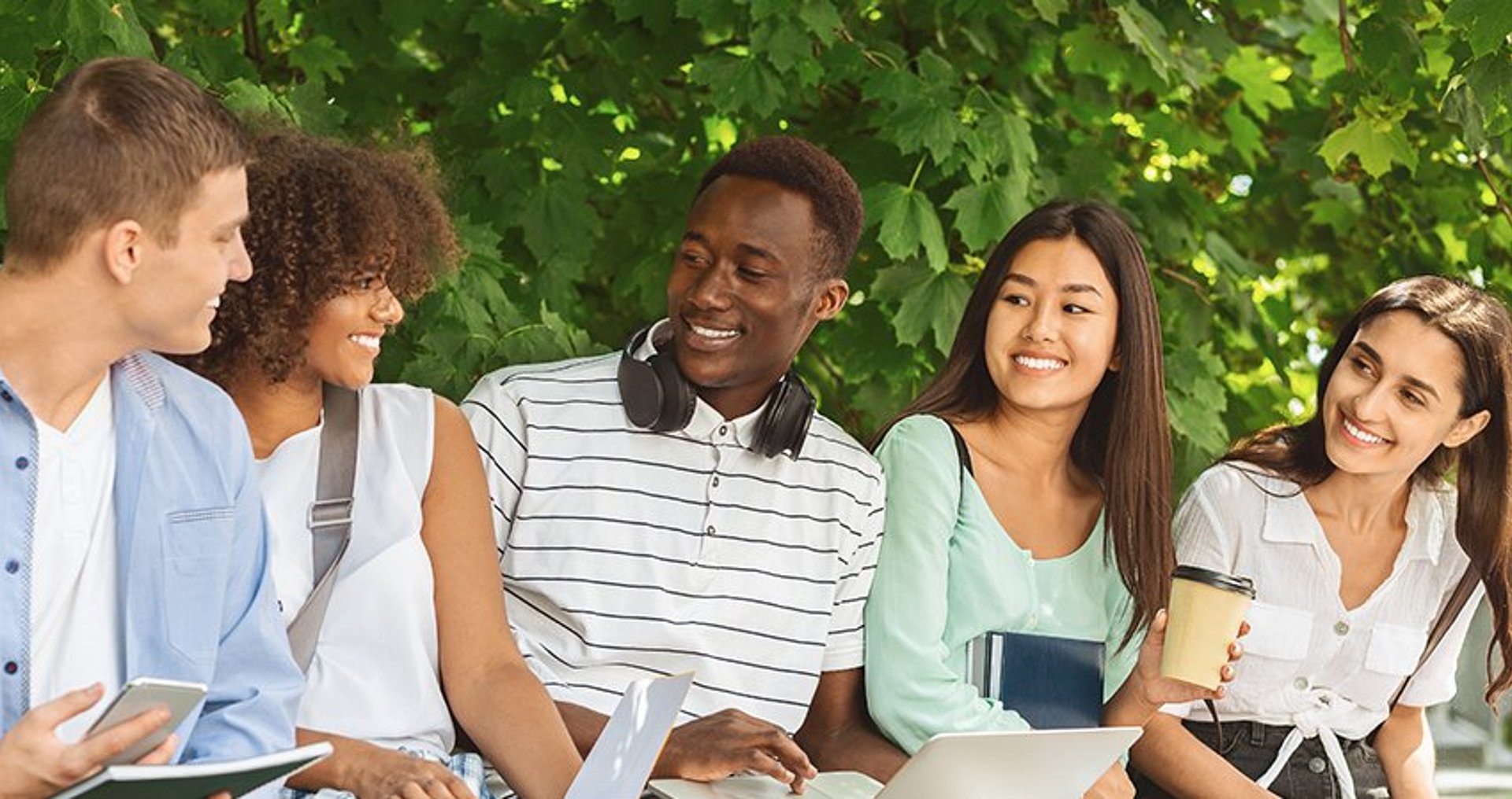 International Students Five students from different backgrounds sit together with their study materials and laptops outdoors against a backdrop of green leaves
