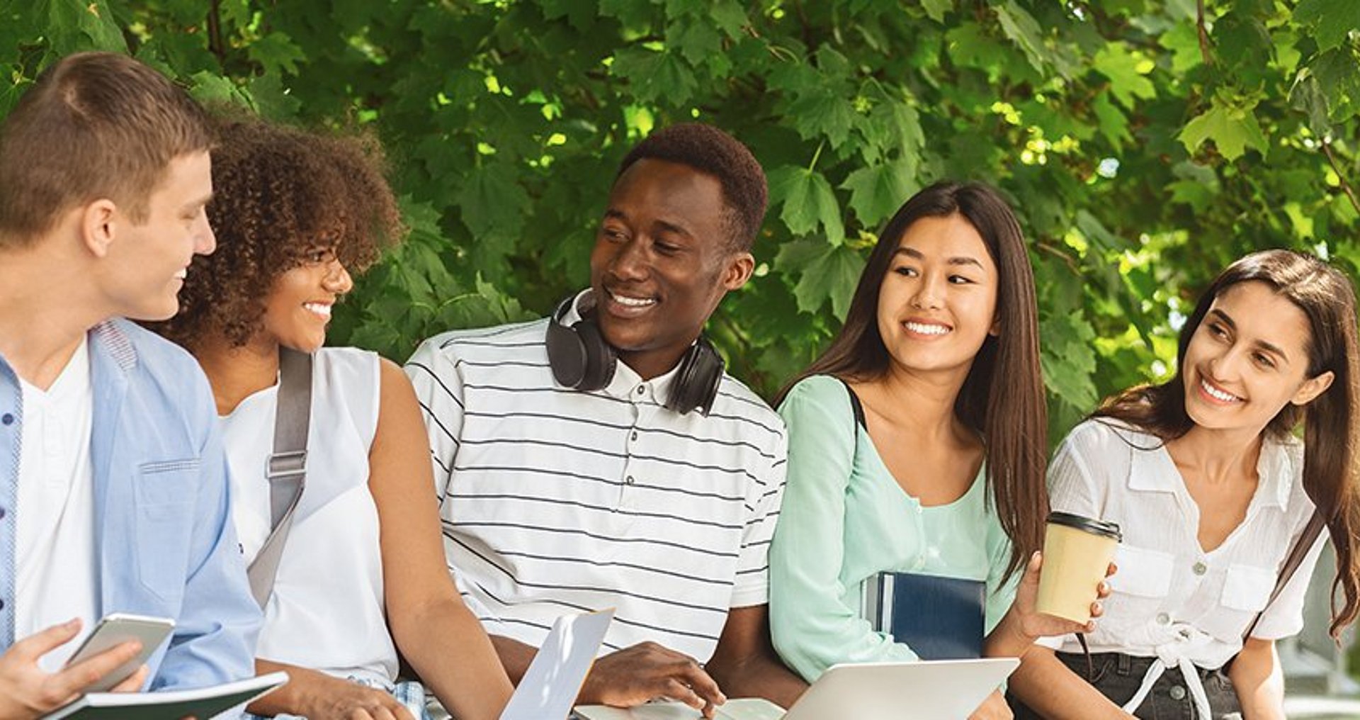Fünf Studierende unterschiedlicher Herkunft sitzen mit ihren Studienunterlagen und Laptops gemeinsam im Freien vor dem Hintergrund grünen Blattwerks