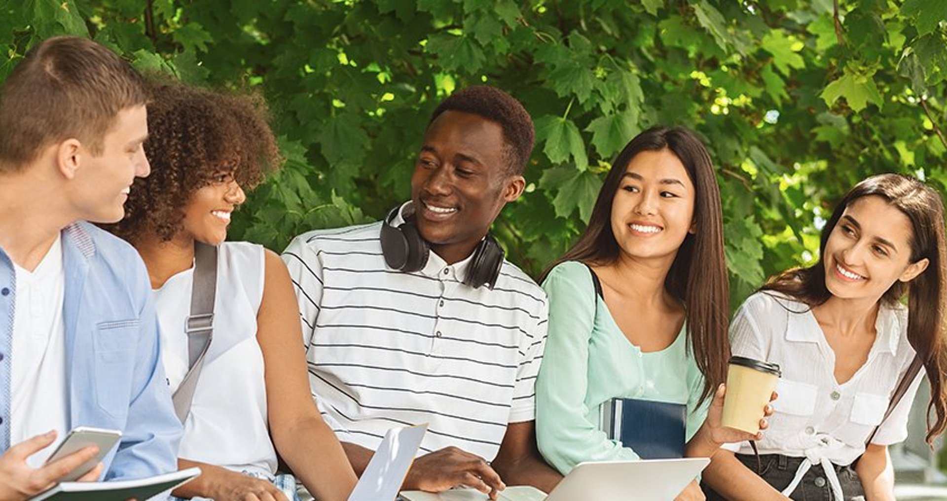 Internationale Studierende Fünf Studierende unterschiedlicher Herkunft sitzen mit ihren Studienunterlagen und Laptops gemeinsam im Freien vor dem Hintergrund grünen Blattwerks