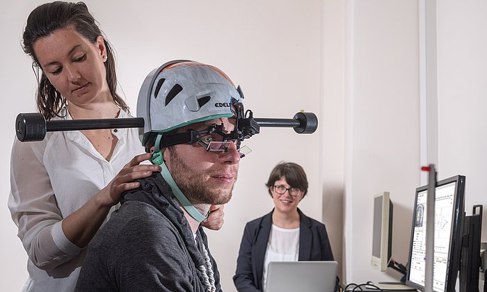 Dr. Cecilia Ramaioli (links) überprüft den Sitz des Helms mit Gewichten bei einem Probanden. Prof. Nadine Lehnen stellt im Hintergrund den Computer für das Experiment ein. 
