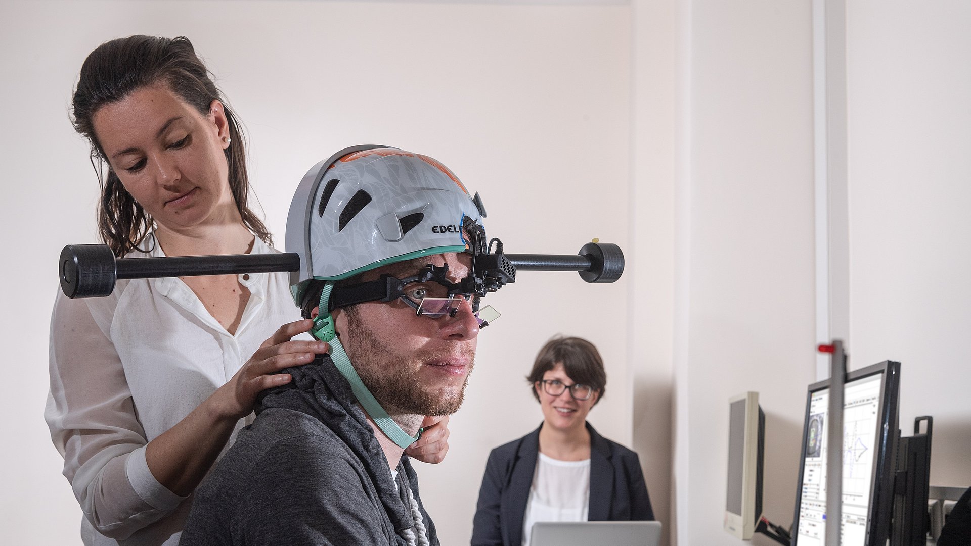 Dr. Cecilia Ramaioli (left) checks if the weighted helmet fits correctly to the head of a study participant. In the background, Prof. Nadine Lehnen prepares the computer for the experiment. 