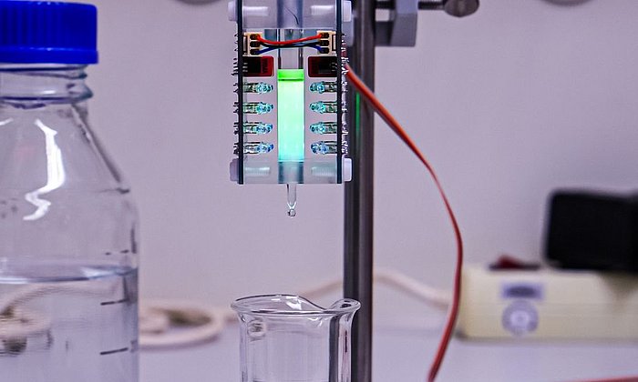 The image shows the apparatus that the team developed for work on a laboratory scale. You can see a small container positioned between LED lights. The liquid in the container glows slightly.
