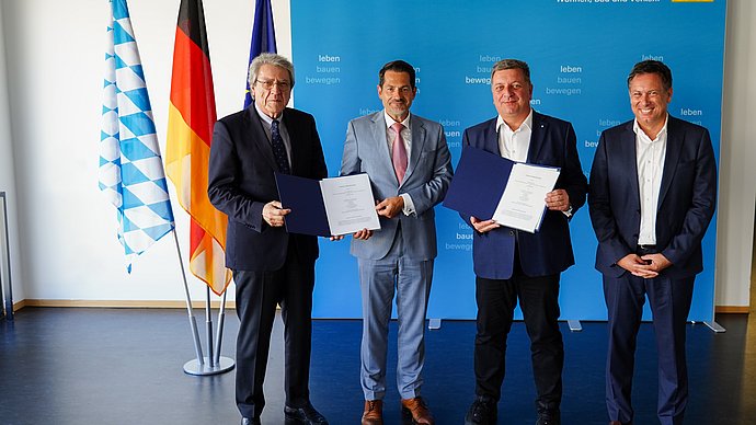 Unterzeichnung der Vereinbarung im Staatsministerium. Vier Männer aus Wissenschaft, Industrie und Politik stehen vor einer Fotowand.