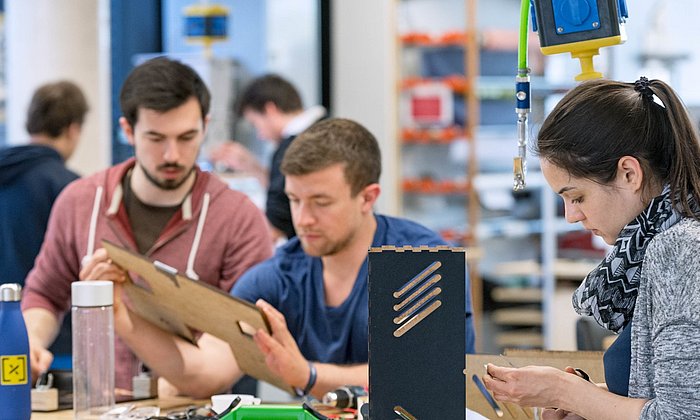 Two male and one female student work together on a model at a table in a workshop.