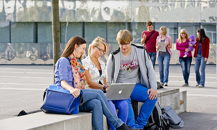 Three students on campus looking into a laptop together. More students in the background.