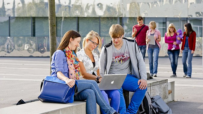 Three students on campus looking into a laptop together. More students in the background.