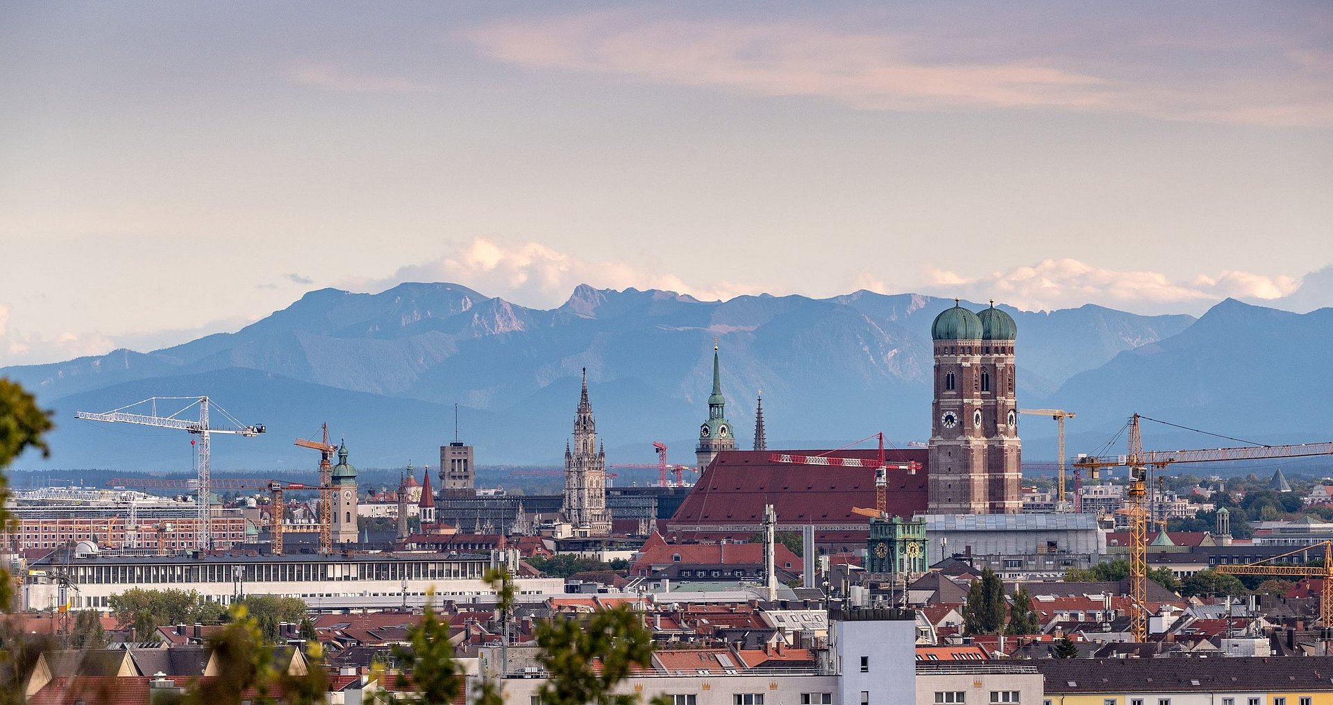 Eine Aufnahme der Stadt München von oben, die Frauenkirche prominent zeigt. Am Horizont sind die Alpen zu sehen.