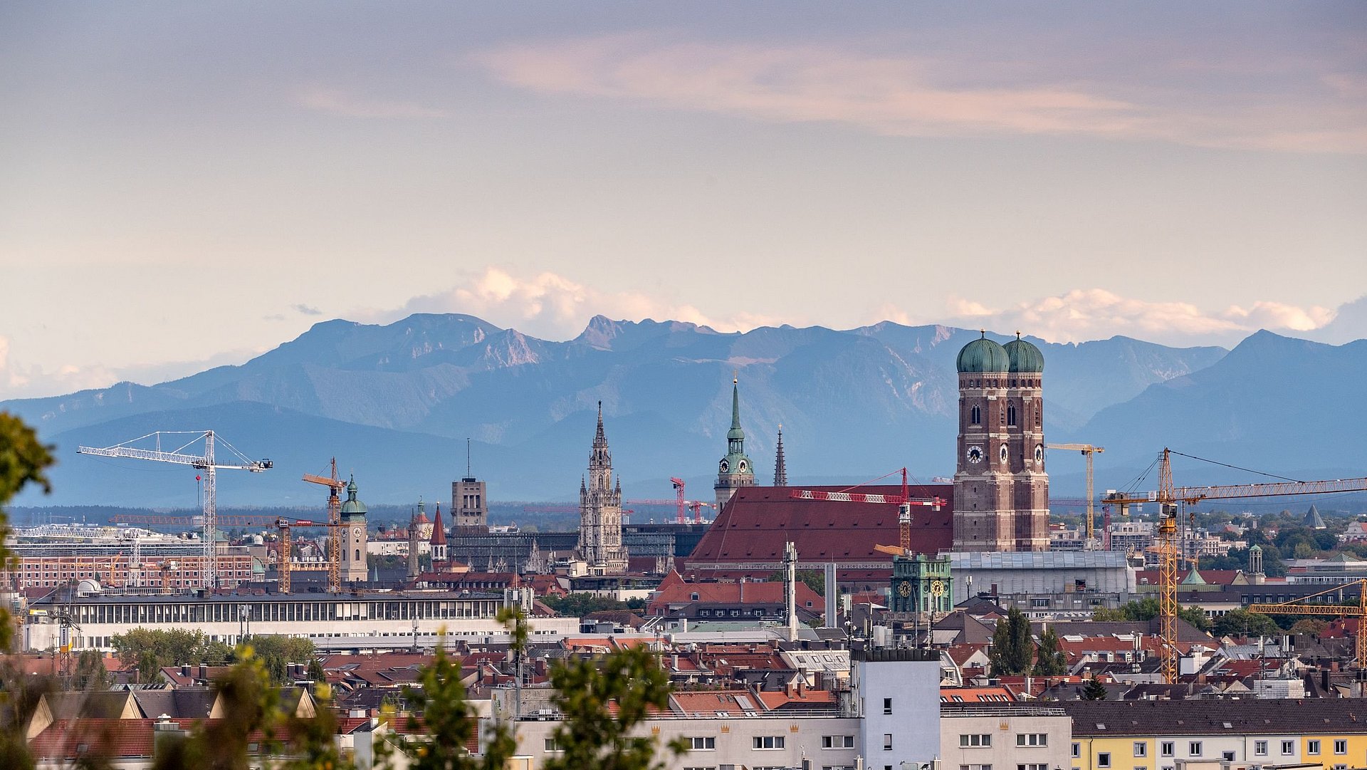 Eine Aufnahme der Stadt München von oben, die Frauenkirche prominent zeigt. Am Horizont sind die Alpen zu sehen.