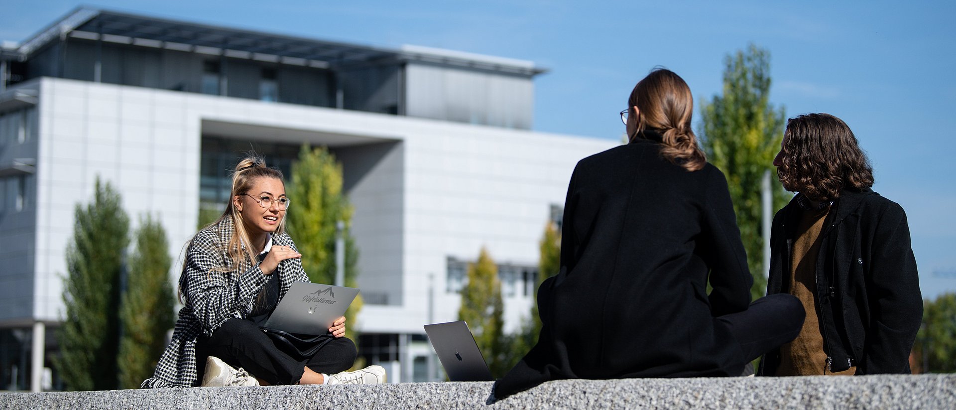 Three female students sit on a stone wall at the Garching campus and talk to each other.