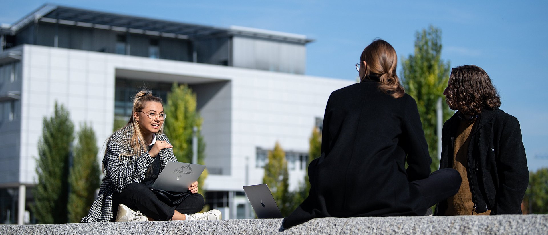 Mit ihren Vorschlägen für den Ideenwettbewerb Academicus können Studierende, Mitarbeitende und Alumni ihren Campus mitgestalten. Drei Studentinnen sitzen auf einer Steinmauer am Campus Garching und sprechen miteinander.