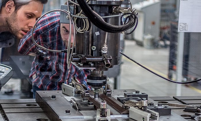 Dr. Andreas Bachmann (li.) beobachtet durch ein Sicherheitsglas, wie der Roboter zwei Metallplatten mit dem schnell drehenden Schweißstift zusammenschweißt.