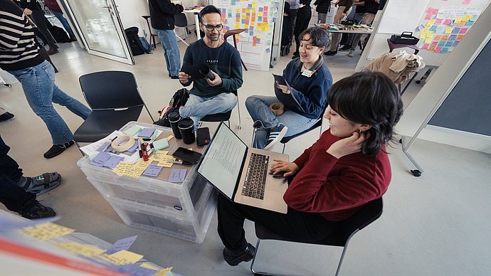 Three students at an improvised table made of two plastic containers, in the background partitions with a multitude of written-on colored sticky notes.