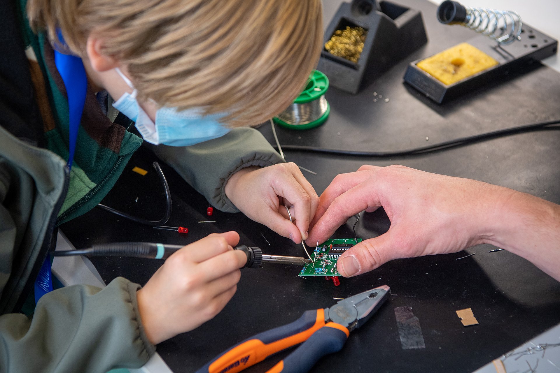 A child wearing a mask and an anorak soldering on a component held by another person.