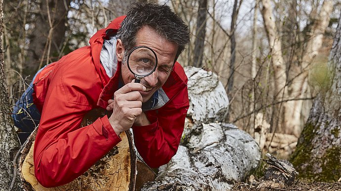 A man is lying on a tree log and looks into the camera, through a magnifying glass. In the background is a forest.