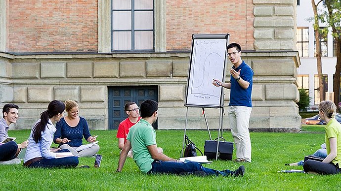 Eine Gruppe Studierender auf einer Wiese vor einem Universitätsgebäude, ein Student präsentiert eine Skizze auf einem Whiteboard