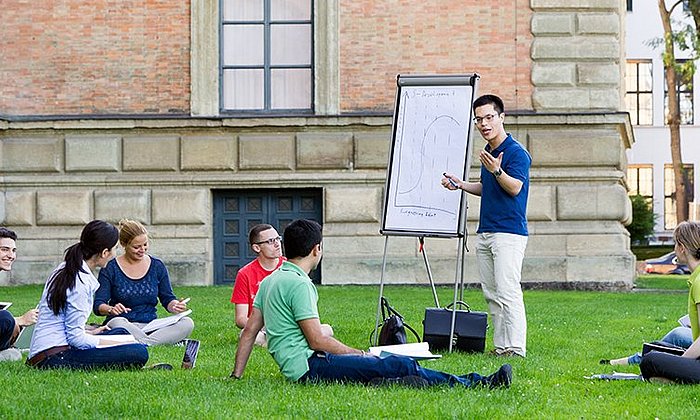 Eine Gruppe Studierender auf einer Wiese vor einem Universitätsgebäude, ein Student präsentiert eine Skizze auf einem Whiteboard