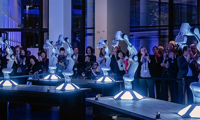 robots are presented in the middle of a big room and people are standing around them taking pictures. The scene is part of the opening event of the Munich School of Robotics and Machine Intelligence in the Pinakothek der Moderne.