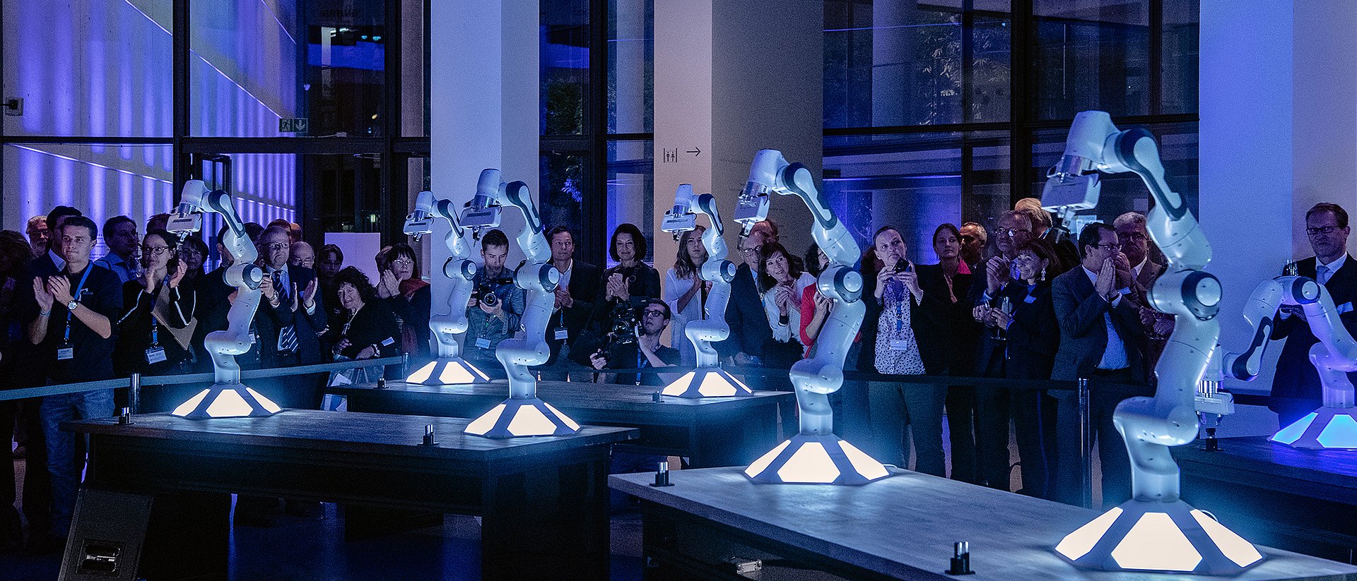 robots are presented in the middle of a big room and people are standing around them taking pictures. The scene is part of the opening event of the Munich School of Robotics and Machine Intelligence in the Pinakothek der Moderne.