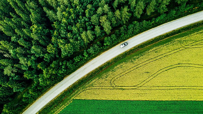 Es ist eine Straße von oben zu sehen, die sich zwischen einem Wald und einem landwirtschaftlichen Feld befindet.
