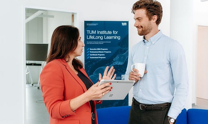 Two people in business attire talking in front of a roll-up of the TUM Institute for LifeLong Learning.