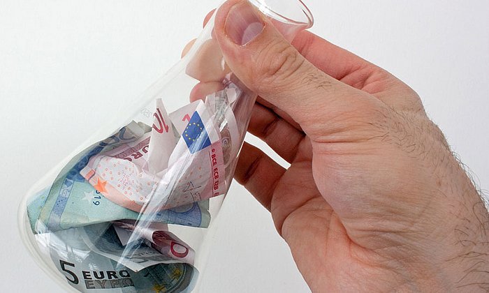 Eine Hand hält ein Reagenzglas gefüllt mit Geldscheinen