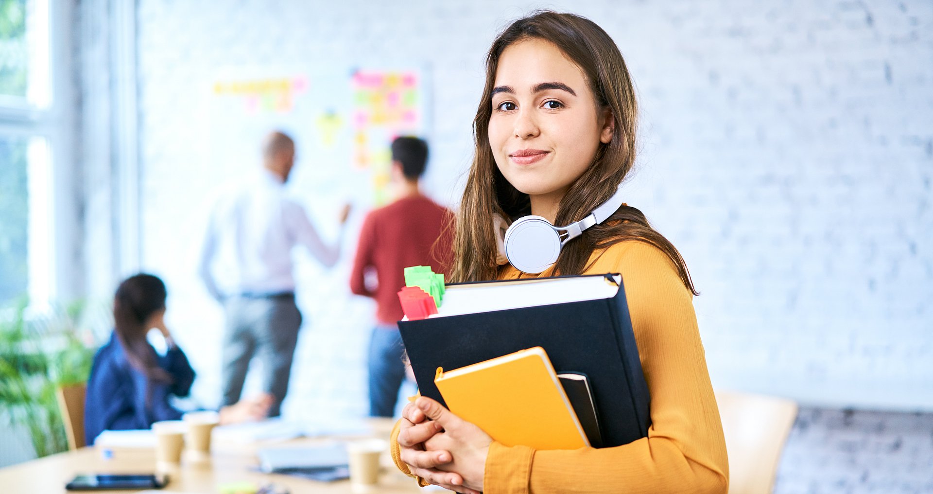 Eine Studentin, die Ordner und Notizbücher festhält, blickt den Betrachter freundlich an; im Hintergrund verschwommen ein Seminarraum, in dem zwei Personen vor einer Wand mit Post-its stehen und miteinander sprechen, während eine dritte Person an einem Tisch im Raum sitzt und zu diesen blickt.