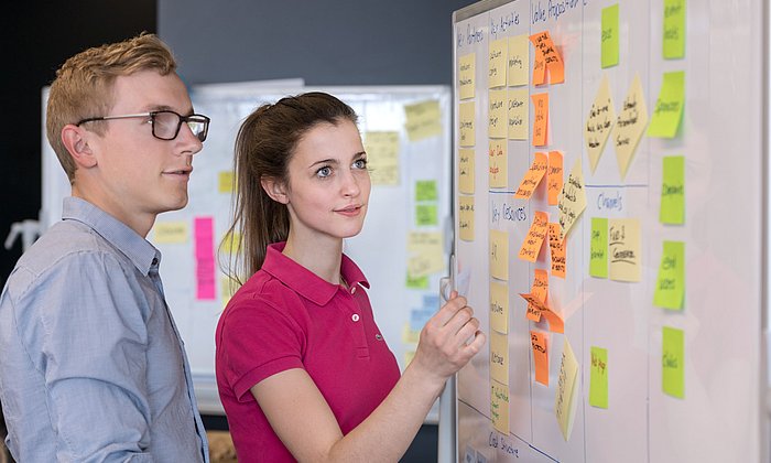 A man and a woman perform the Kanban method on a whiteboard