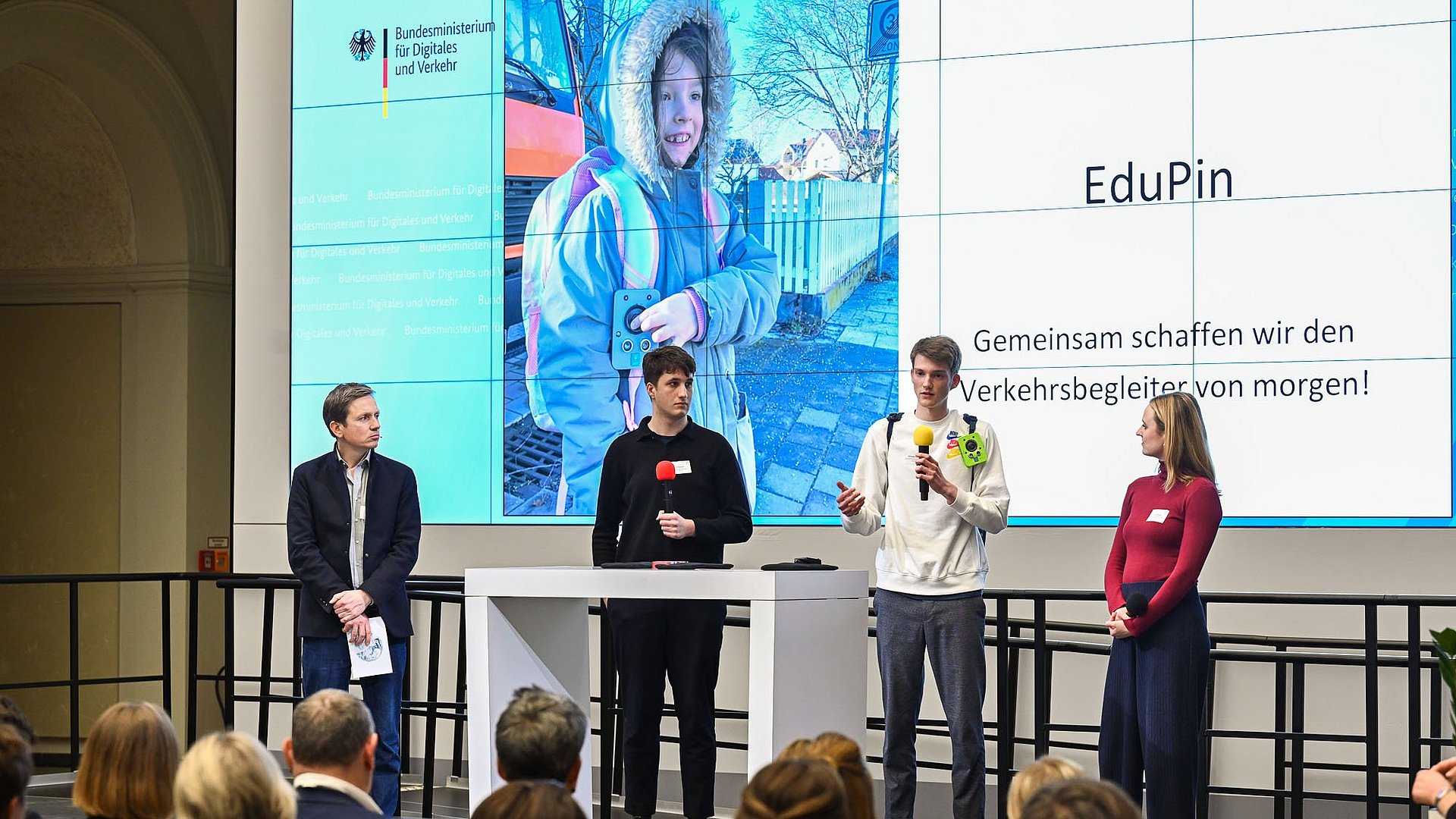 Team EduPin presents its concept for child-friendly transportation planning for Digital and Transport presenting their concept to the audience, behind them a presentation projected onto the wall showing a child wearing a prototype of the EduPin on the strap of his backpack and handling it, as well as the words "EduPin. Together we are creating the transportation companion of tomorrow!".
