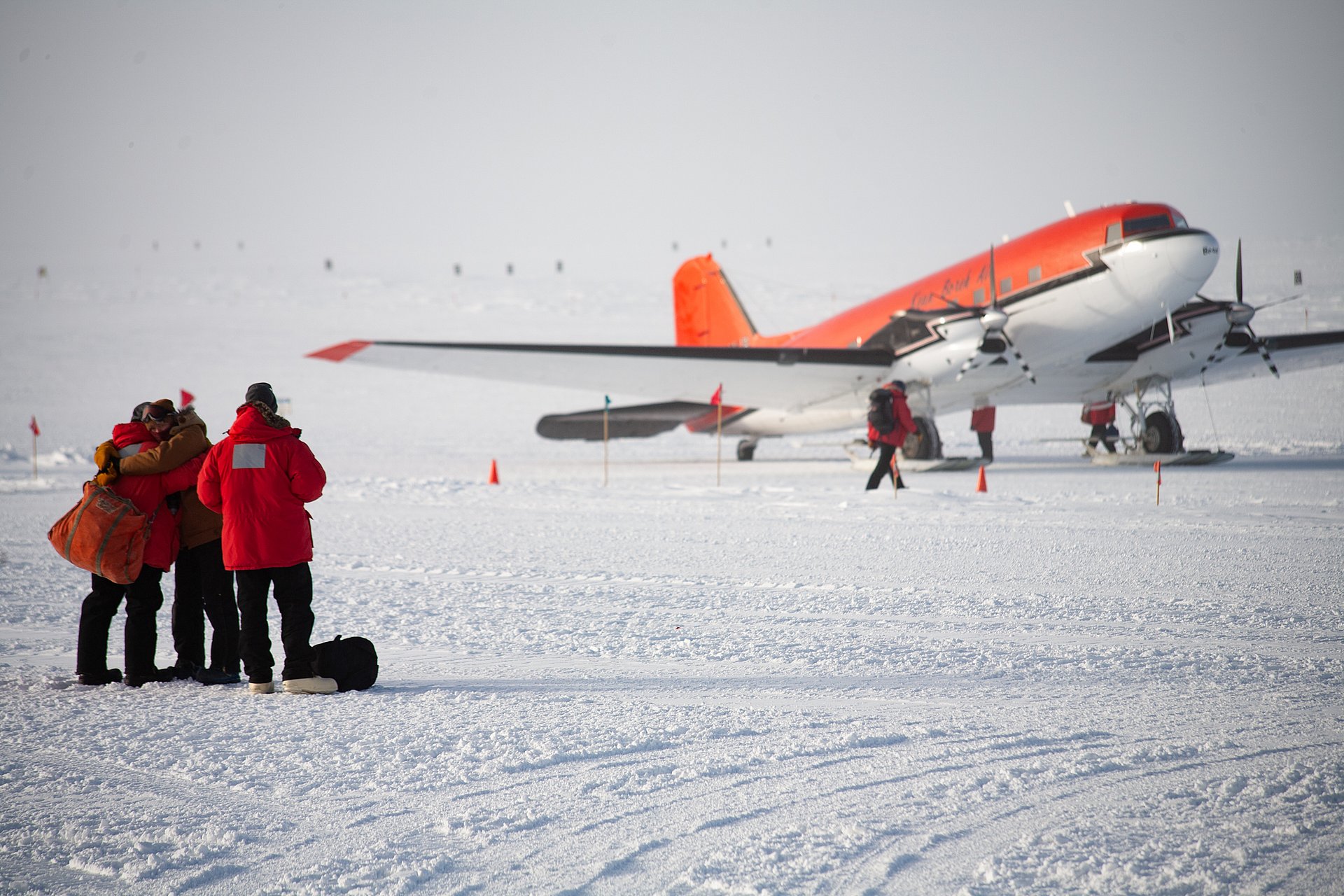 Im Hintergrund steht ein Passagierflugzeug im Schnee, im Vordergrund eine Gruppe warm angezogener Menschen, die sich verabschieden. 