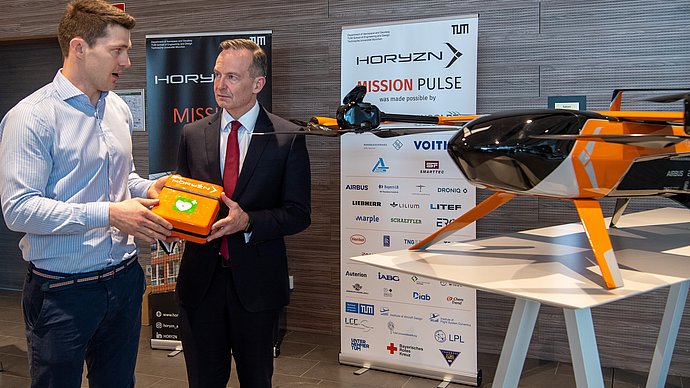 Zwei Personen halten eine Box vor einer Drohne hoch, in der ein Defibrillator zu findne ist. 