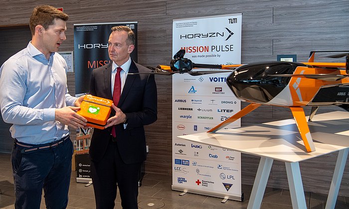 Zwei Personen halten eine Box vor einer Drohne hoch, in der ein Defibrillator zu findne ist. 