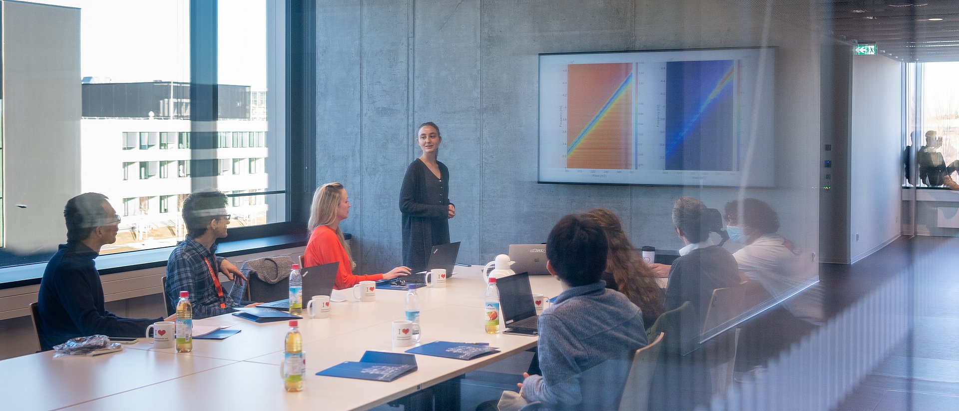 A woman leads through a presentation in a seminar room photographed through a pane of glass. She is standing at a conference table where 7 other people are sitting. A graphic is projected on the wall.