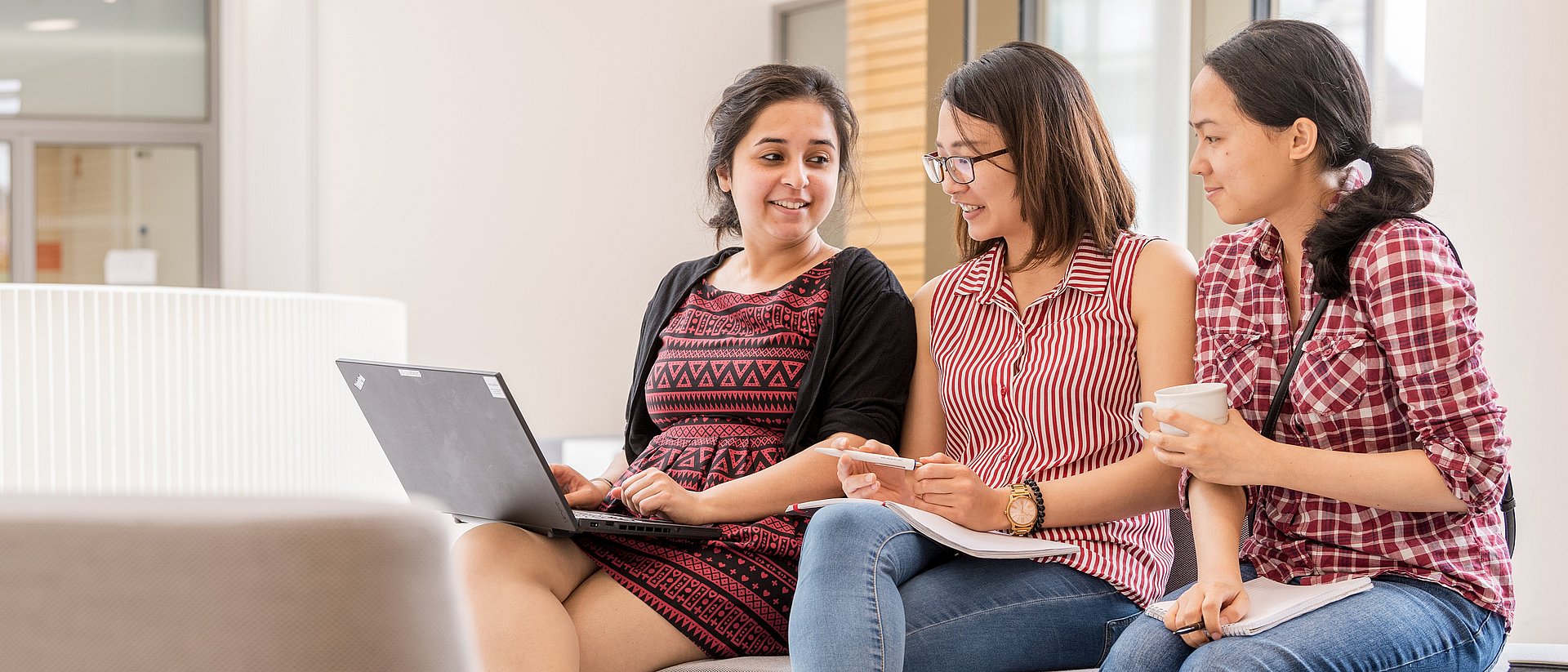 Drei Studentinnen sitzen zusammen mit einem Laptop und unterhalten sich. 