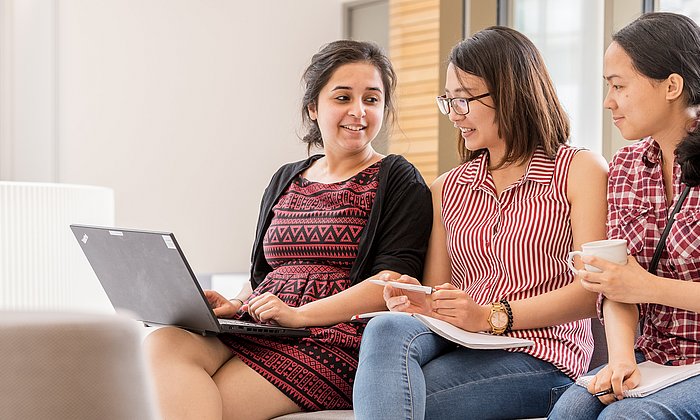 Drei Studentinnen sitzen zusammen mit einem Laptop und unterhalten sich. 