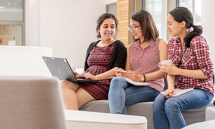 Drei Studentinnen sitzen zusammen mit einem Laptop und unterhalten sich. 