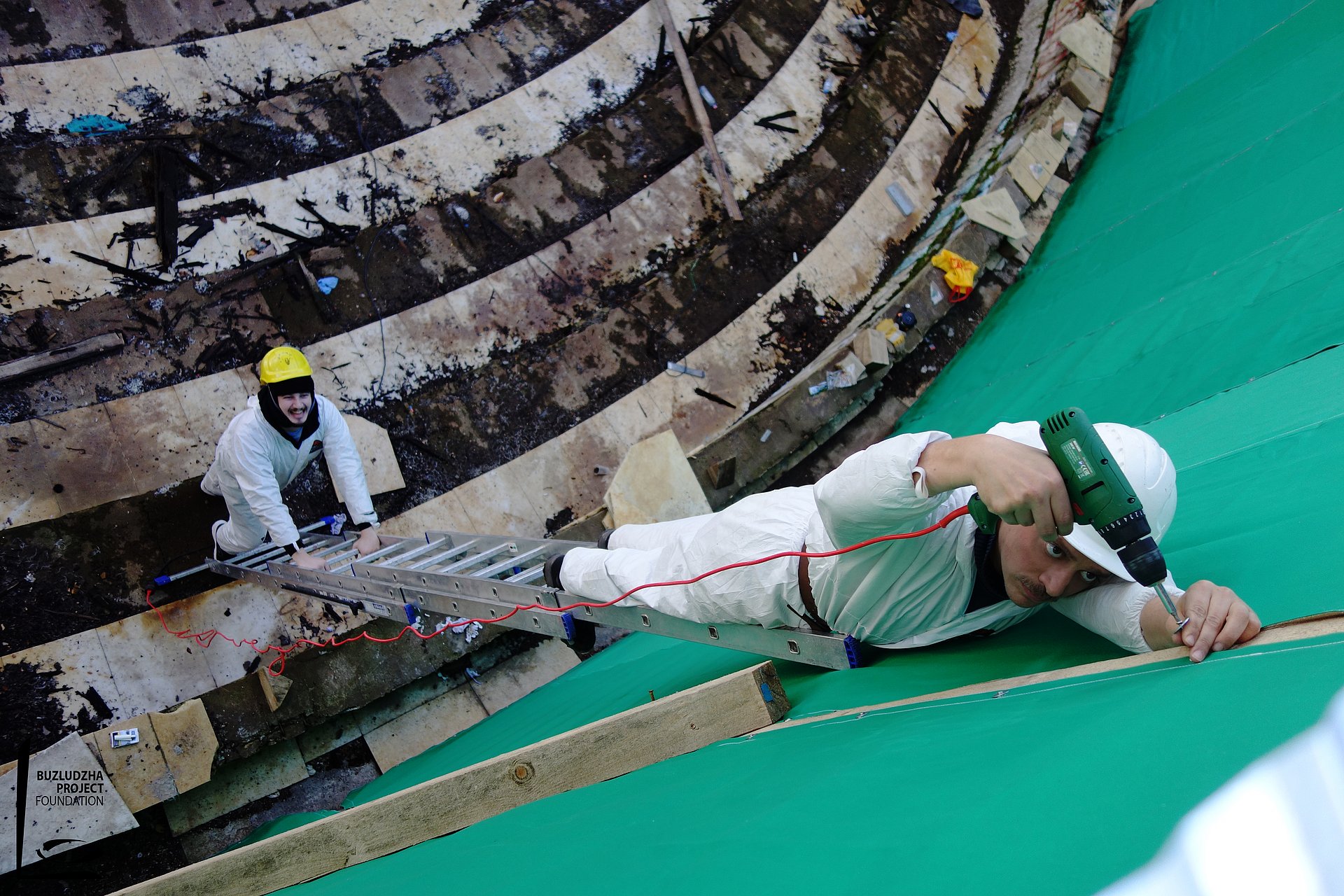 A man stands on a ladder in protective suit and drives a screw with machine into the inner wall of the dome of the Buzludzha monument. Another man in protective suit holds the ladder.