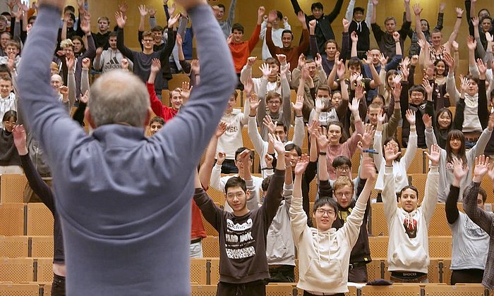 Activity break during the lecture: Prof. Diepold can be seen in the lecture hall from behind, stretching his arms upwards, students in front of him, also with their arms stretched upwards