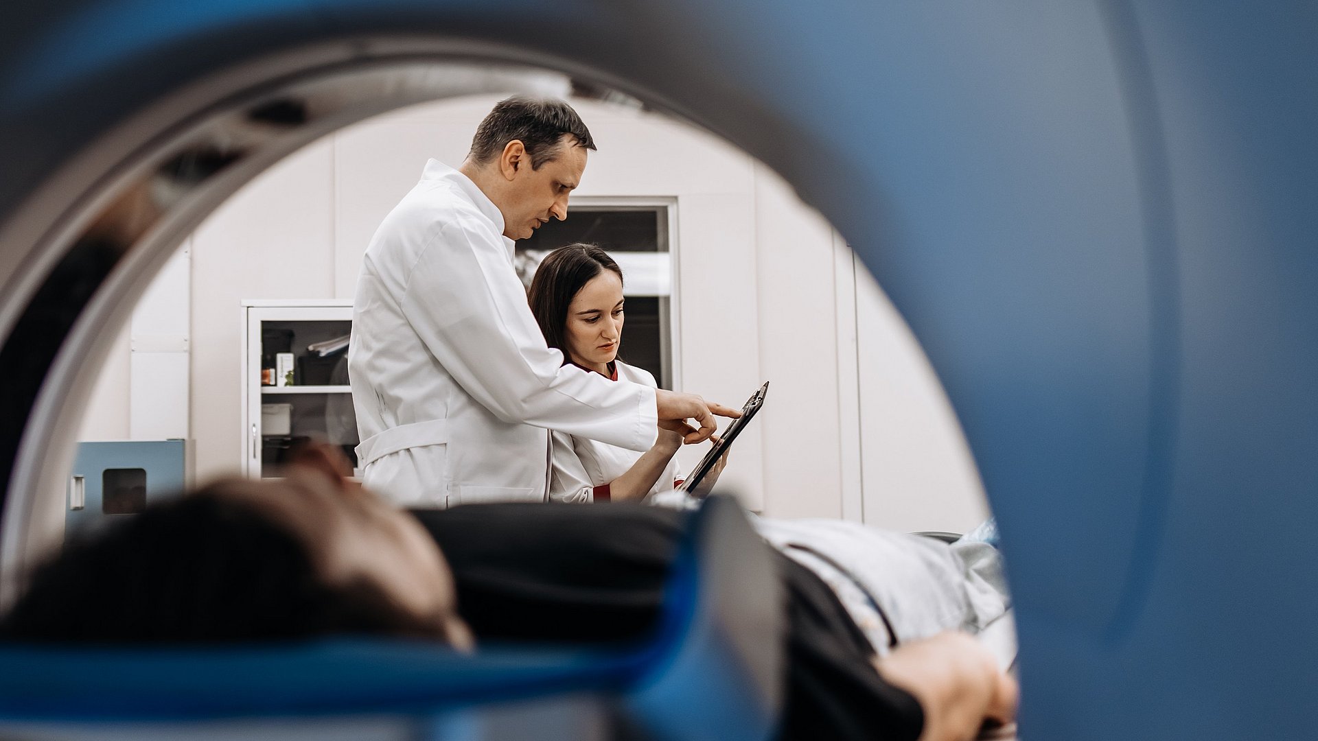 View of a patient and two doctors through an MRI machine. View of a patient and two doctors through an MRI machine.