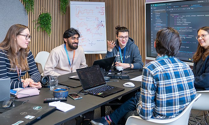 Fünf Studierende sitzen um einen Tisch und diskutieren. Vor zwei Studierenden steht ein Laptop. Sowohl auf den Laptops als auch auf dem Bildschirm im Hintergrund ist Quellcode zu sehen.
