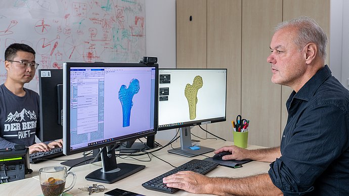 Zwei Forschende vor ihren Computern werden dargestellt. Auf den Bildschirmen sind zwei Hüftimplantate abgebildet, die mithilfe unterschiedlicher Ansätze konstruiert wurden.