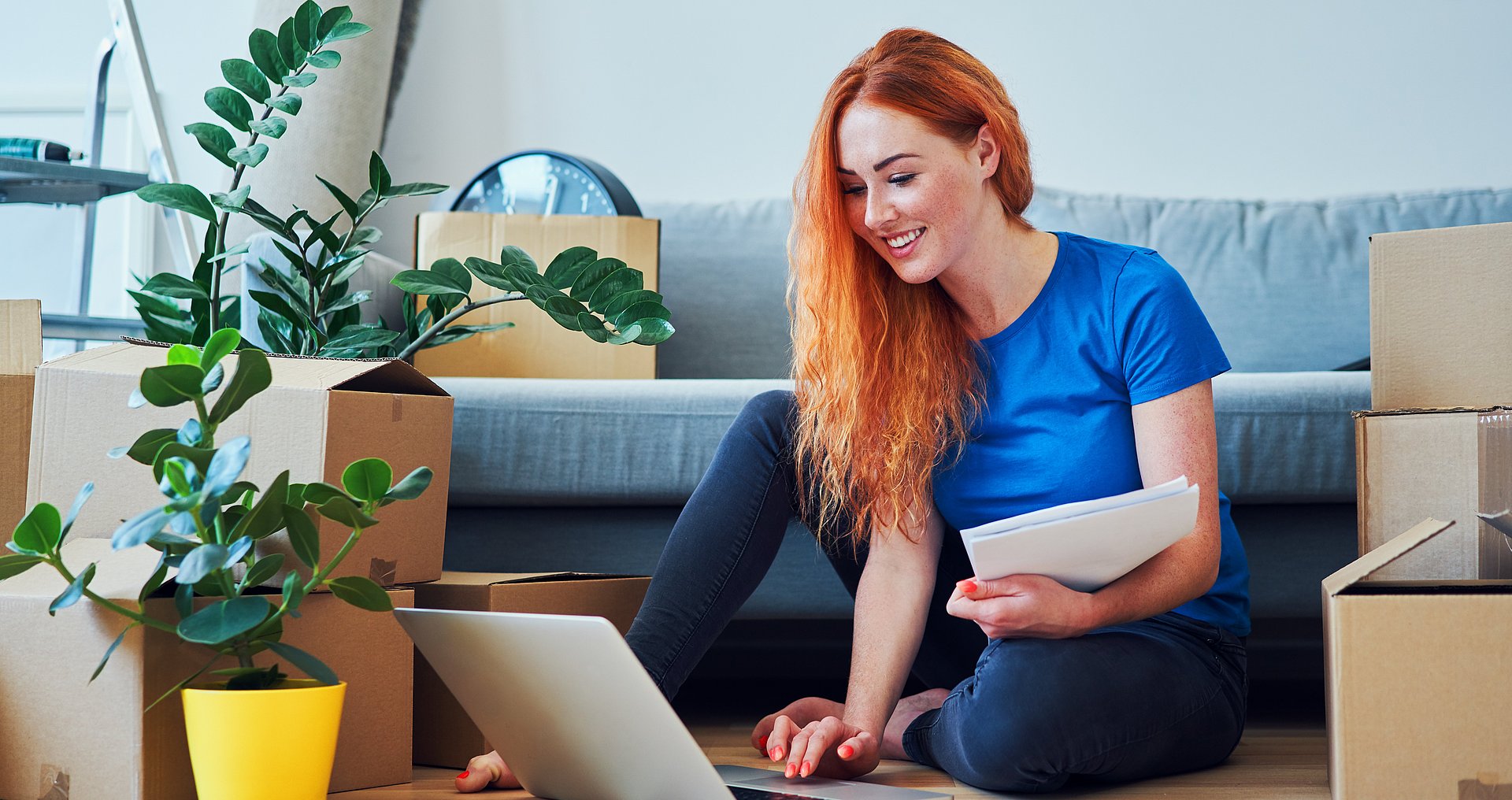 Wohnen und arbeiten © baranq / Adobe Stock Eine junge Frau sitzt in einer Wohnung auf dem Boden am ihrem Laptop, im Hintergrund ein Sofa, darauf und daneben noch nicht ausgepackte Umzugskisten.