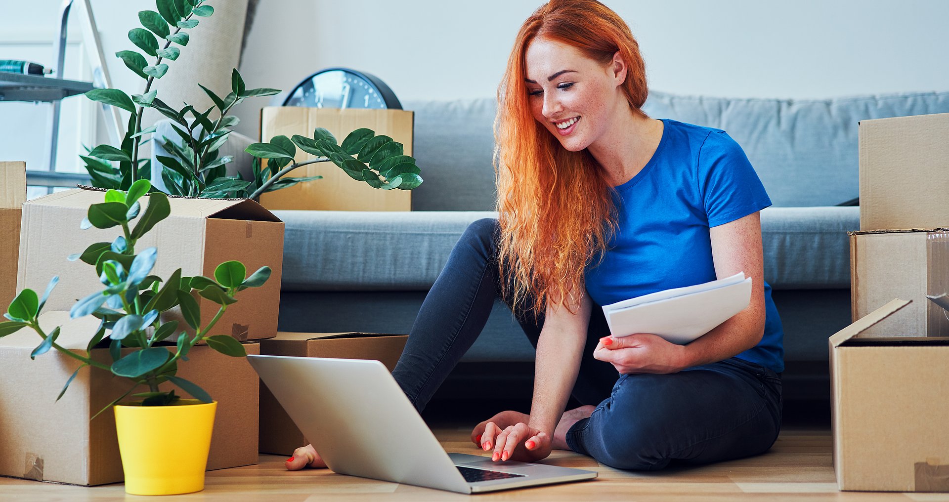 A young woman sits in an apartment on the floor at her laptop, in the background a sofa, on it and next to it not yet unpacked moving boxes.