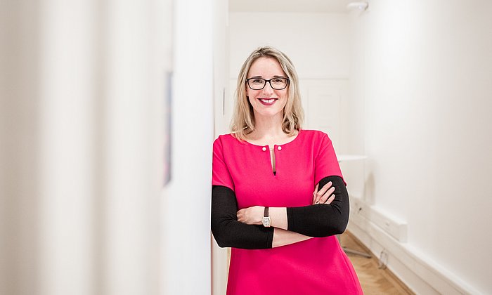 A blond woman is standing in a corridor smiling directly to the camera