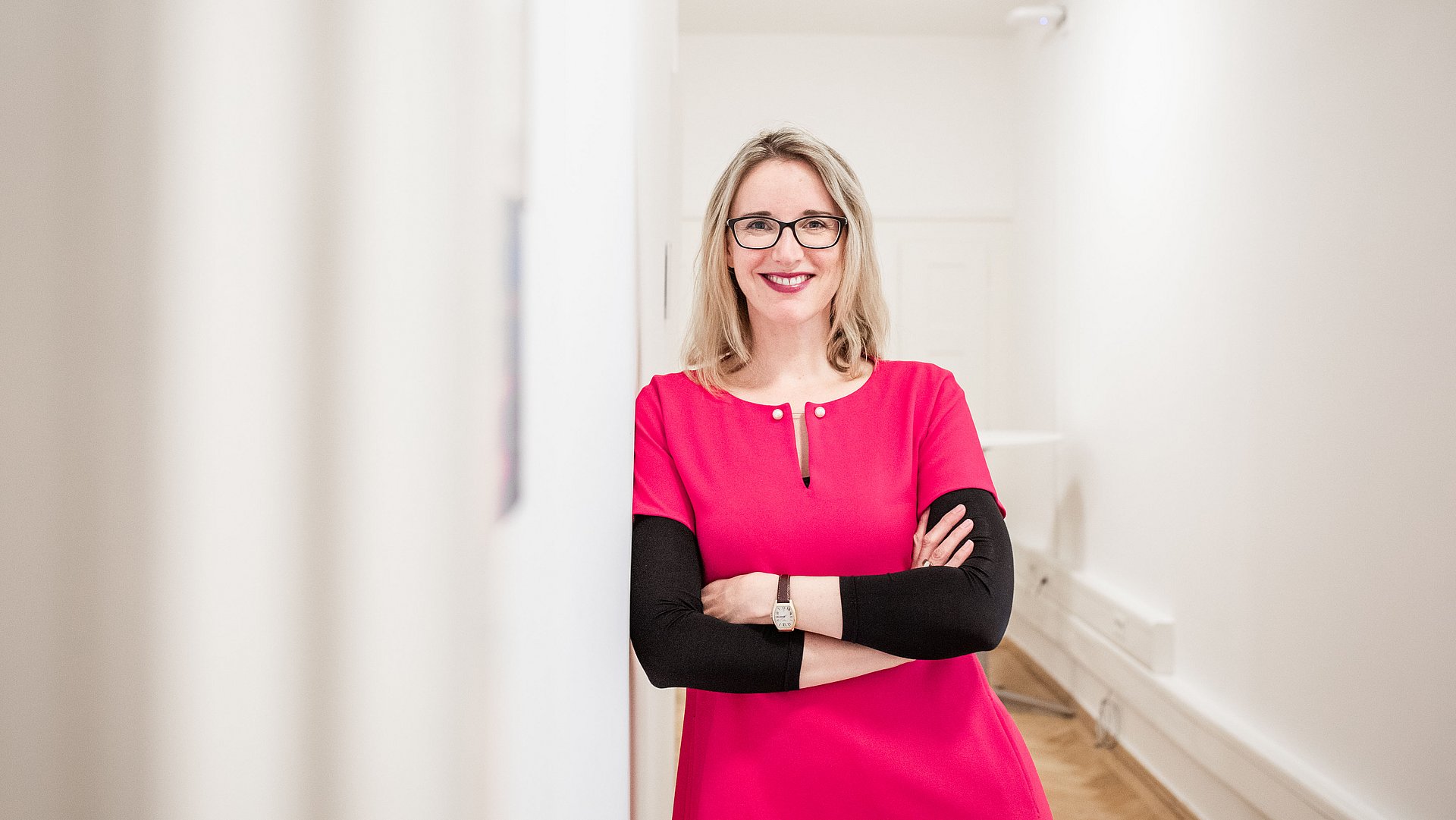 A blond woman is standing in a corridor smiling directly to the camera