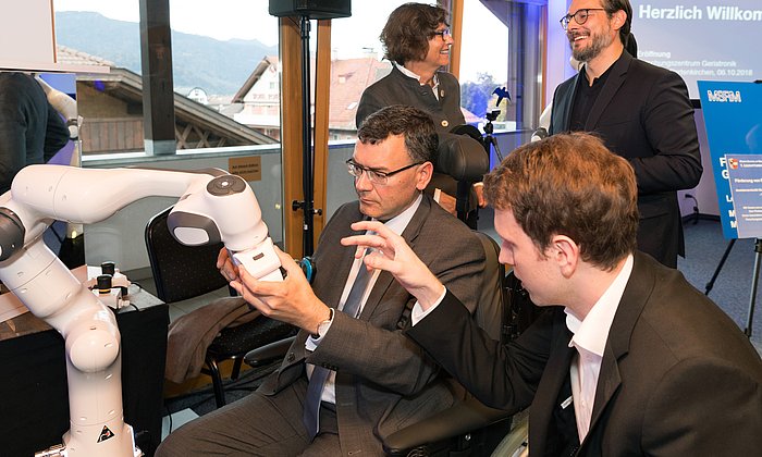 Bavarian State Mnister Dr. Florian Hermann tests a shaving robot. In the background, Dr. Sigrid Meierhofer, mayor of Garmisch-Partenkirchen and Professor Sami Haddadin are talking.