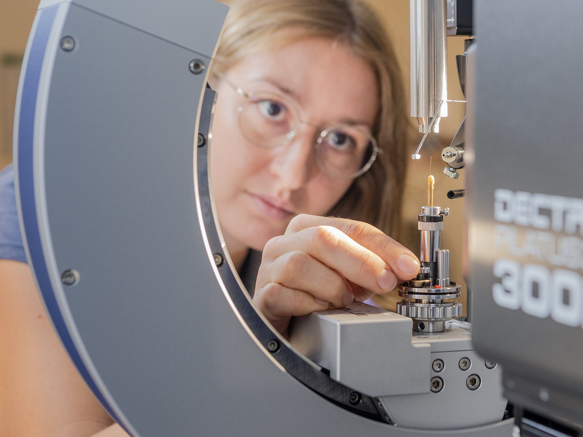 Anna Vogel mounting a single crystal on the single crystal diffractometer. In front of her, a glass capillary can be seen on which a crystal of about 0.2 mm diameter is located. Data are recorded with suitable measuring radiation in order to determine the structure from this.